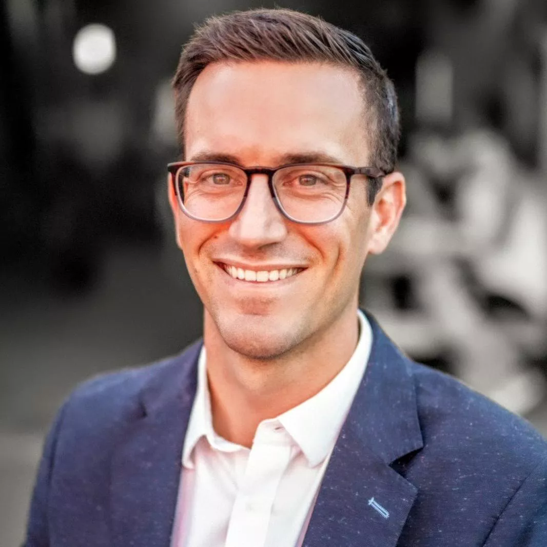 A man wearing a white shirt and a blue blazer is captured in an outdoor setting. The background features a blurred urban or event ambiance with soft lighting. The image conveys a professional and polished style.